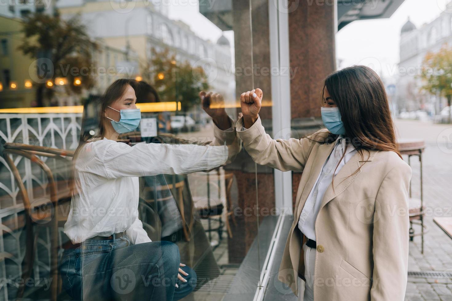 Two women in protective masks opposite each other, window between them. photo