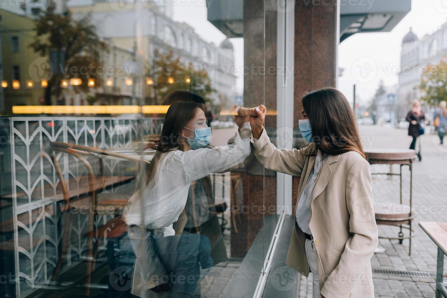 Two women in protective masks opposite each other, window between them. photo
