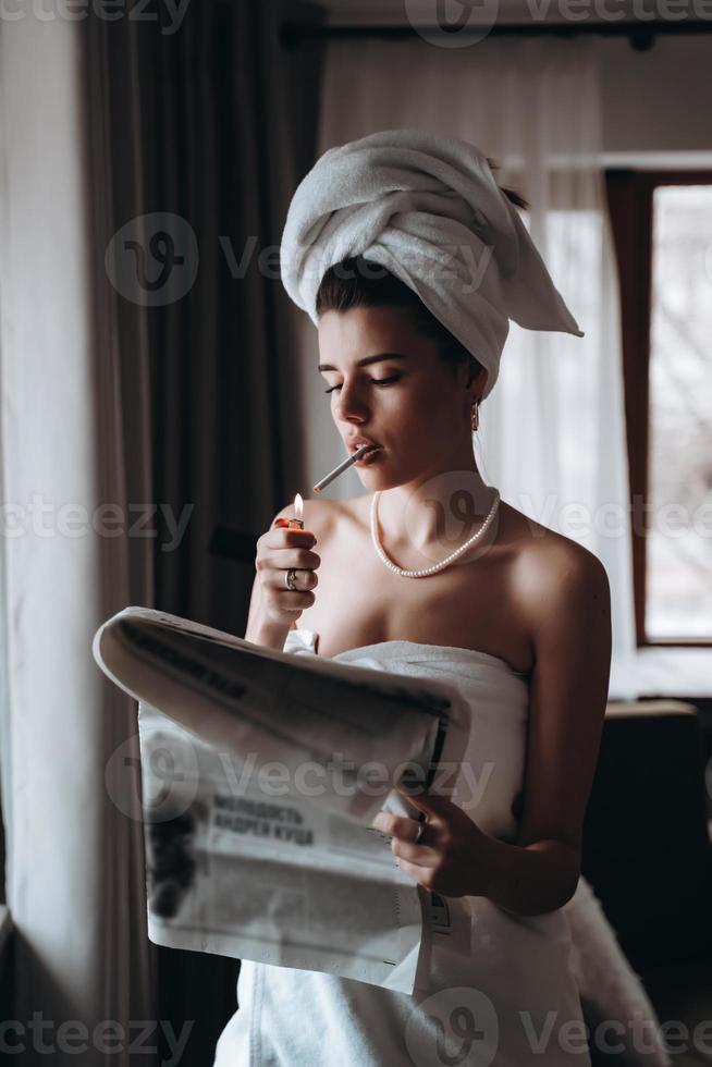 hermosa joven en una toalla fuma un cigarrillo y lee el periódico foto