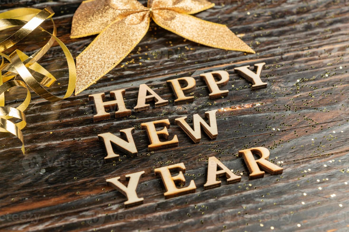 Happy new year wooden taxt on dark wooden background with golden tied bow and scattered confetti. New year greeting card. photo