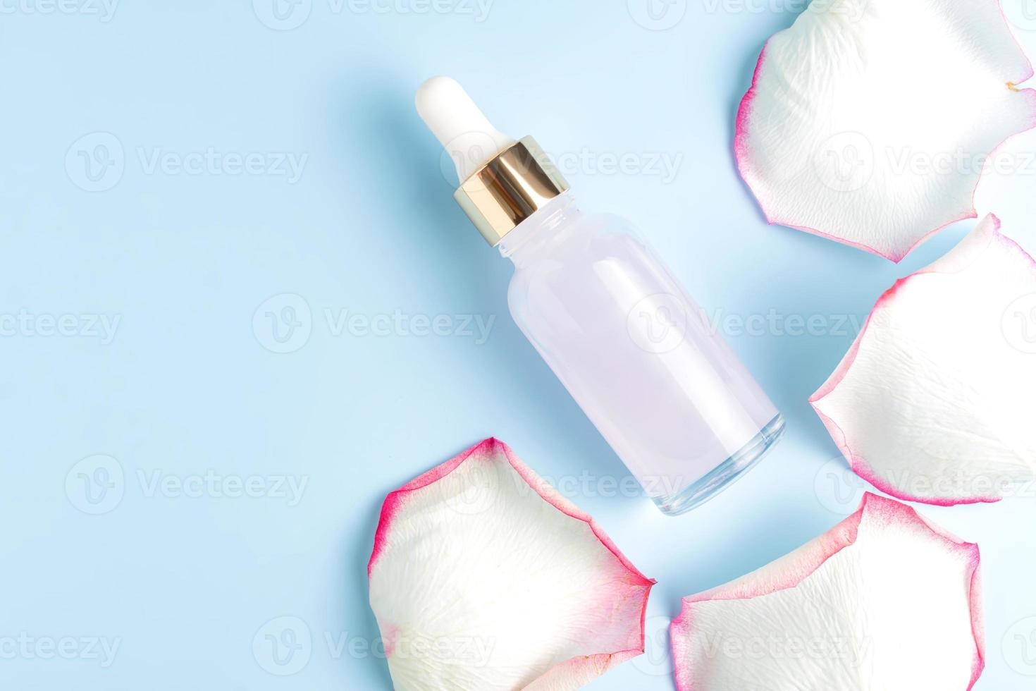 A transparent glass bottle with collagen or hyaluronic acid on a blue background with a rose scattered petals. Place for text. Cosmetic, skin care serum, gentle care. photo