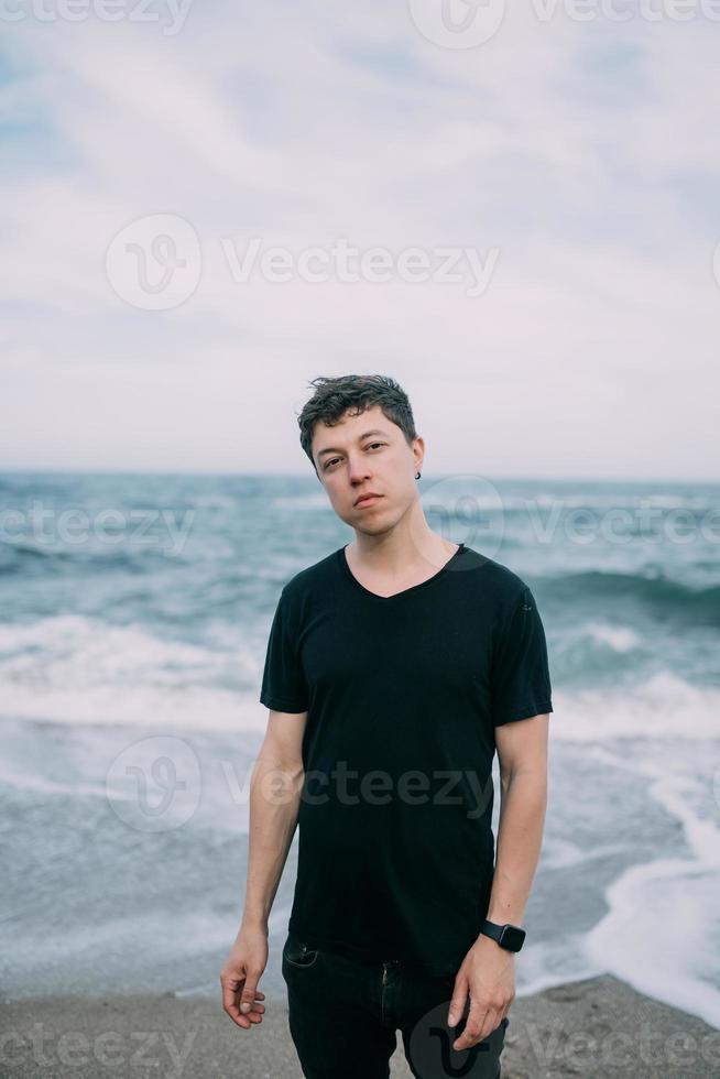 Smiling guy in a black T-shirt stands on the sandy seashore. photo