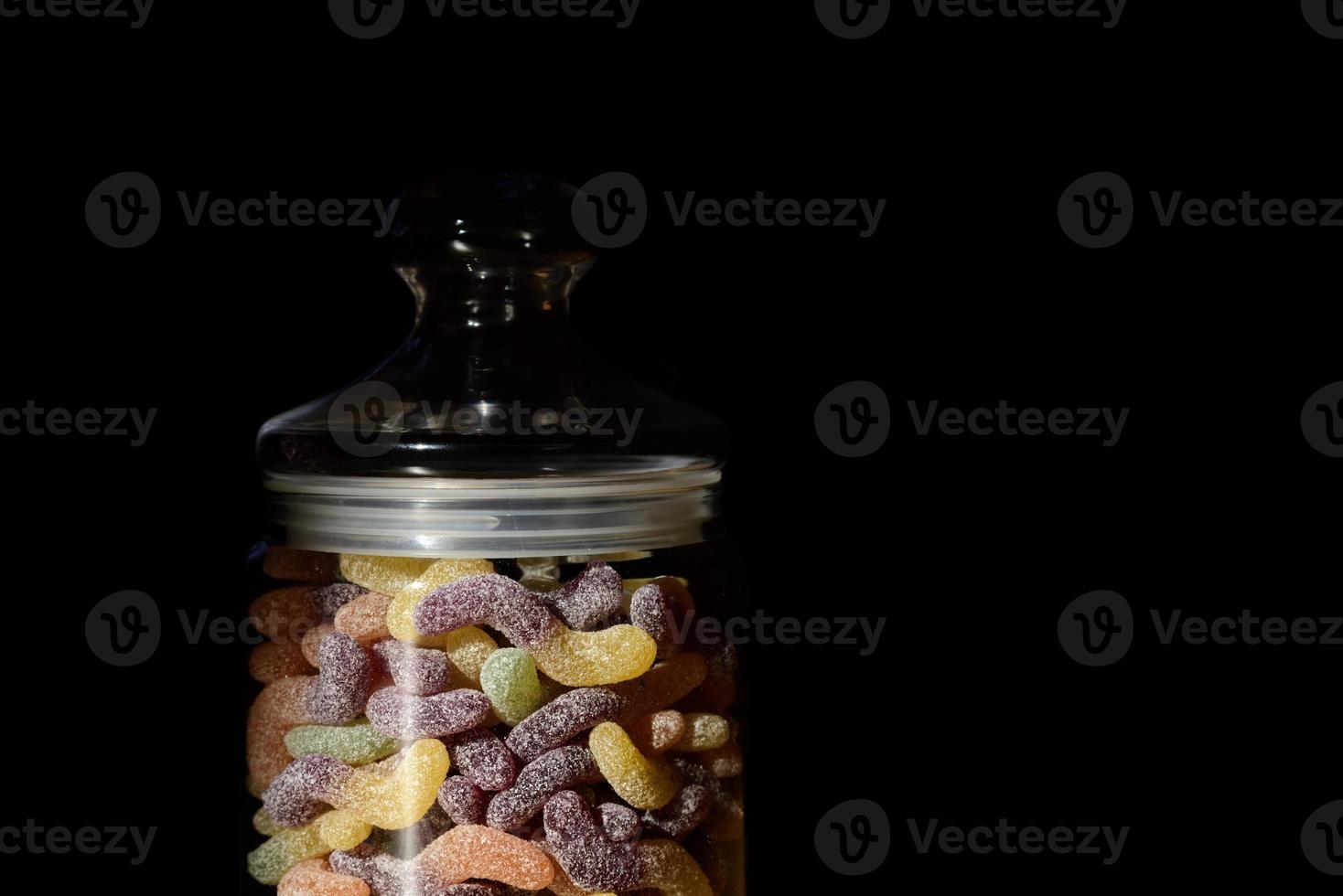 A jar with a lid contains colorful sweets in the form of colorful, sugared soft gums, against a dark background photo