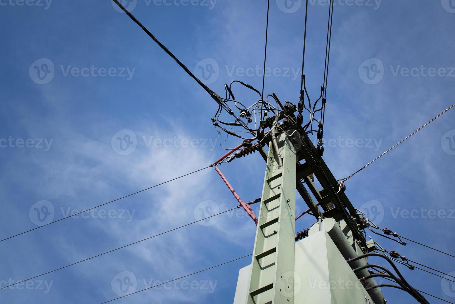 A mast with a network of many different power cables is photographed from below against the blue sky with clouds photo