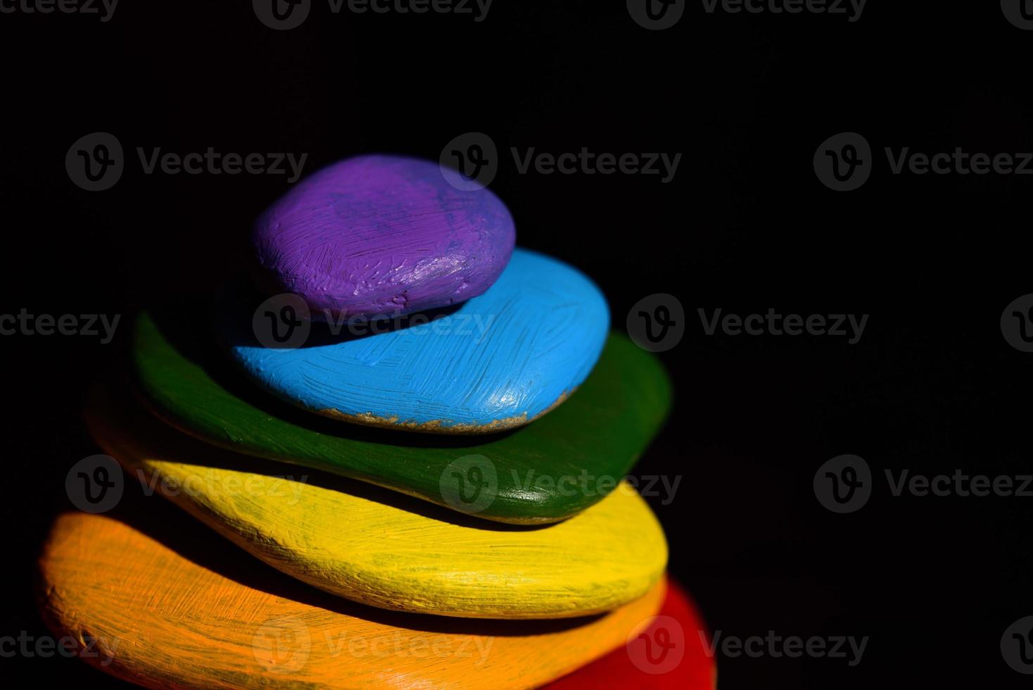 Several brightly colored pebbles painted in rainbow colors lie on top of one another in front of a dark background photo