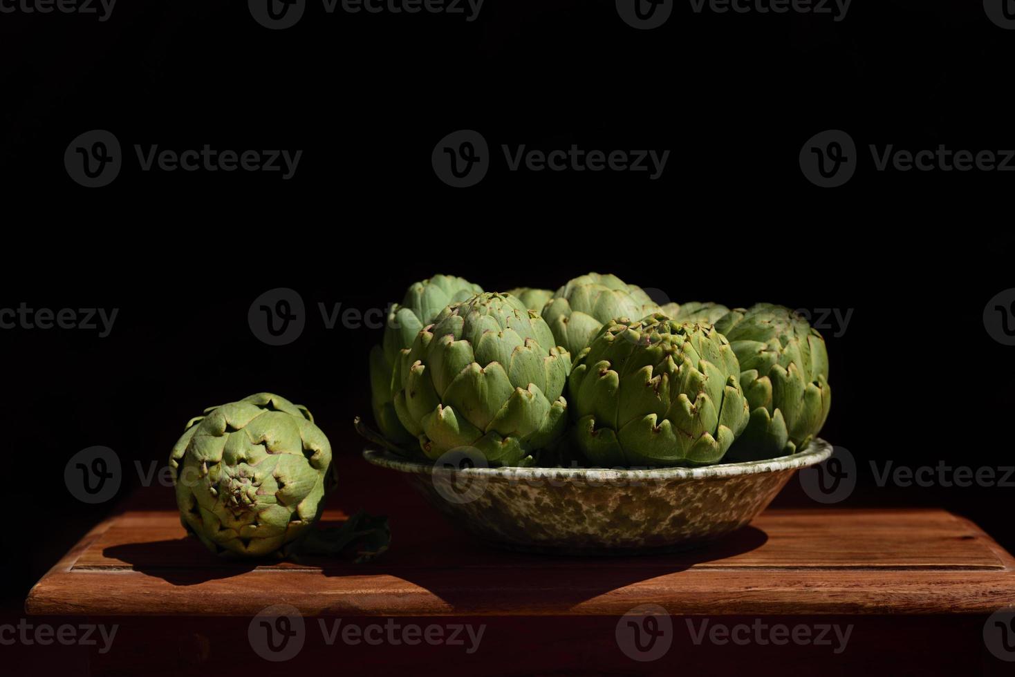 Several arichokes lie in a plate on a wooden table, against a dark background photo