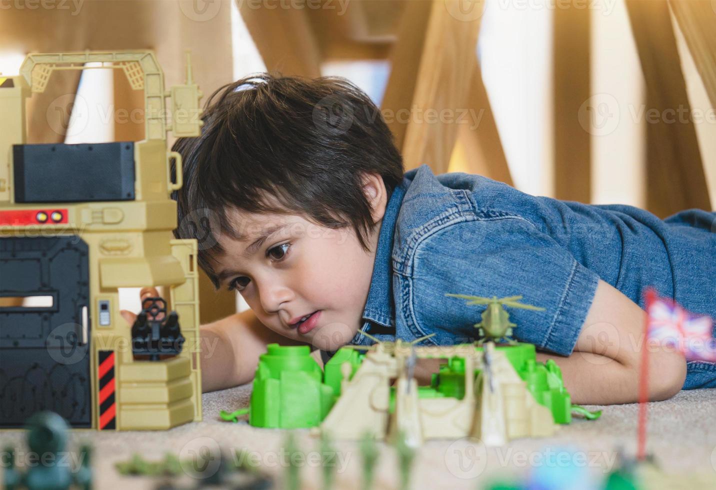 Portrait of School boy lying down on the carpet floor playing with soldiers, military car and figurine toys, Happy Kid playing wars and peace on his own at home, Children imagination and development photo