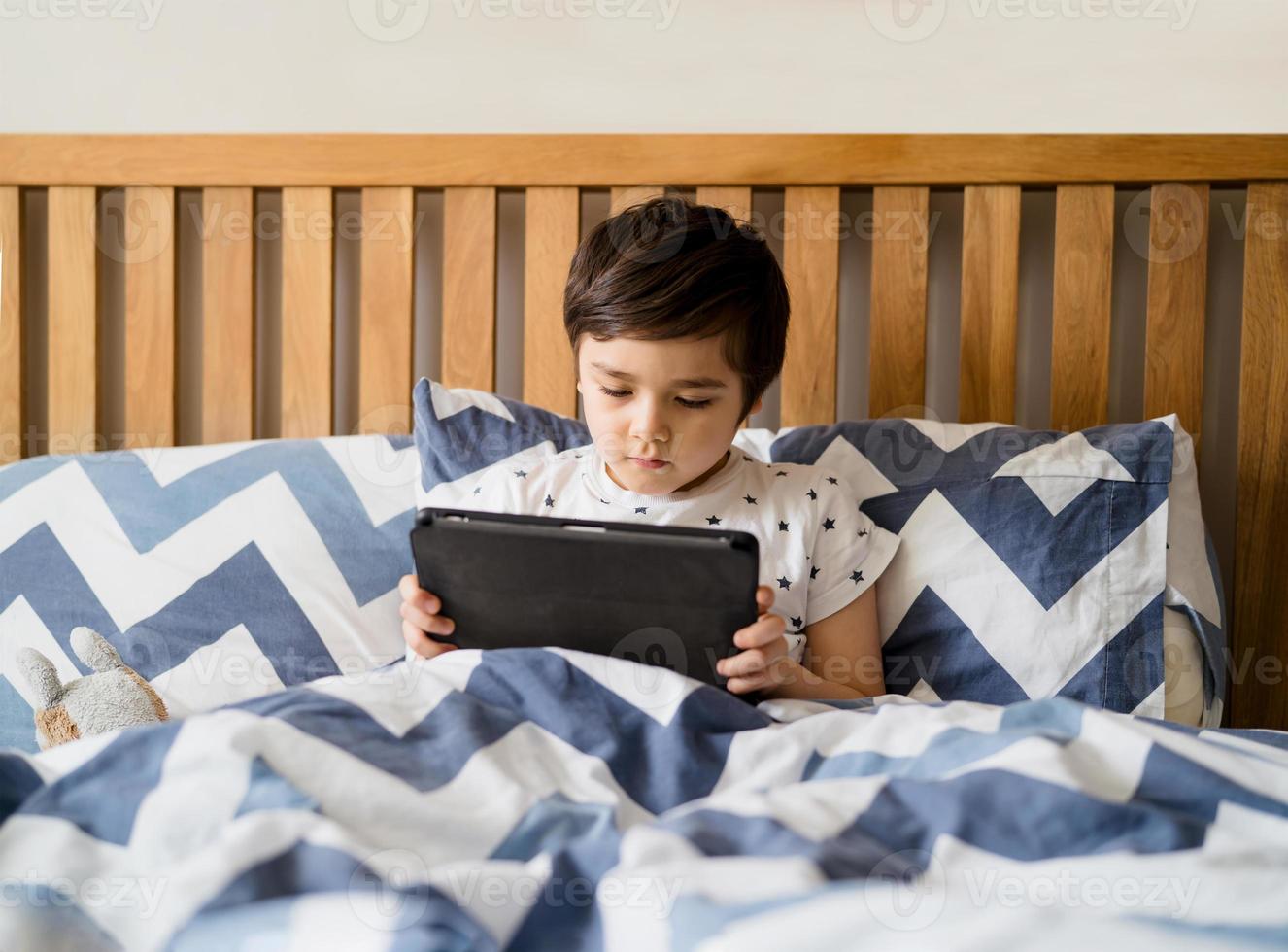 Kid reading bed time stories on tablet before sleep, Happy boy sitting in bed playing games on digital pad, Child relaxing at home in his bed room on weekend. photo