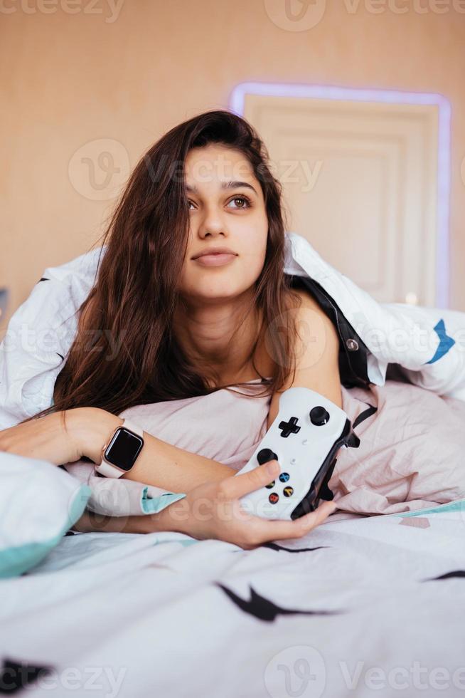 chica divertida acostada en la cama y jugando videojuegos, sosteniendo el controlador foto