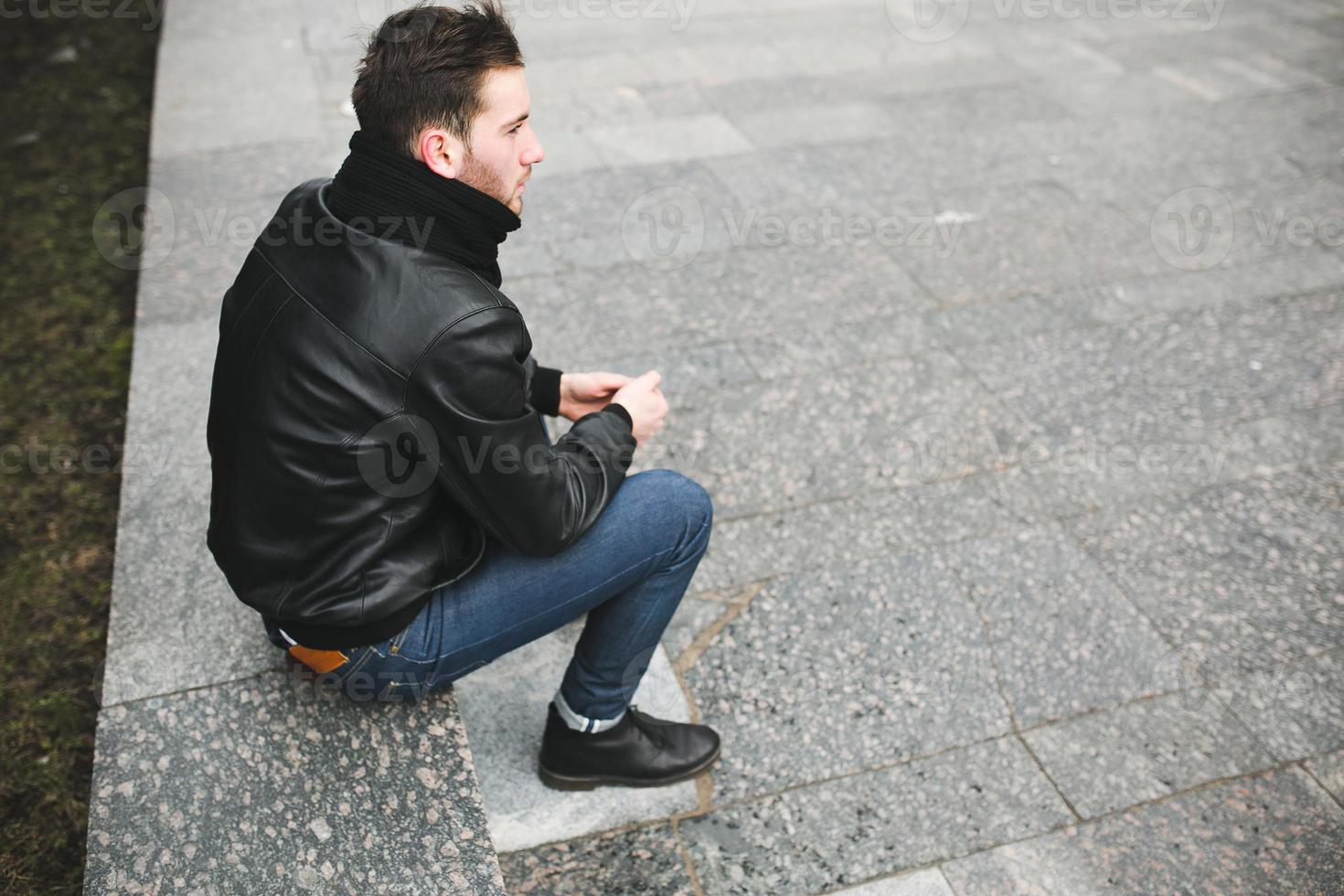 A man dressed in jeans and black jacket seats on a slab photo