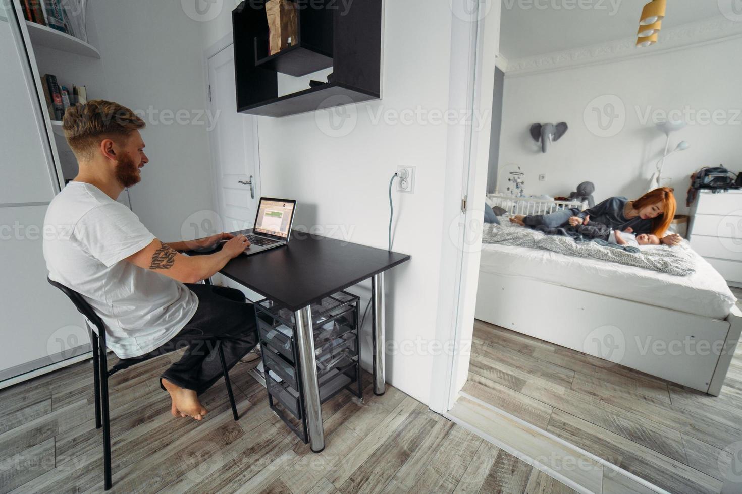 Dad works at laptop, mom and baby on the bed photo