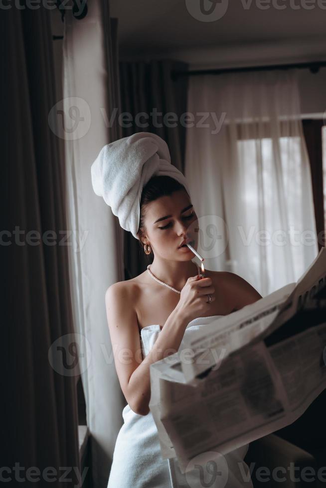 hermosa joven en una toalla fuma un cigarrillo y lee el periódico foto