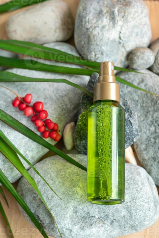 A bottle of green color with a spray bottle lies on the stones, next to the green leaves and berries of schisandra chinensis photo