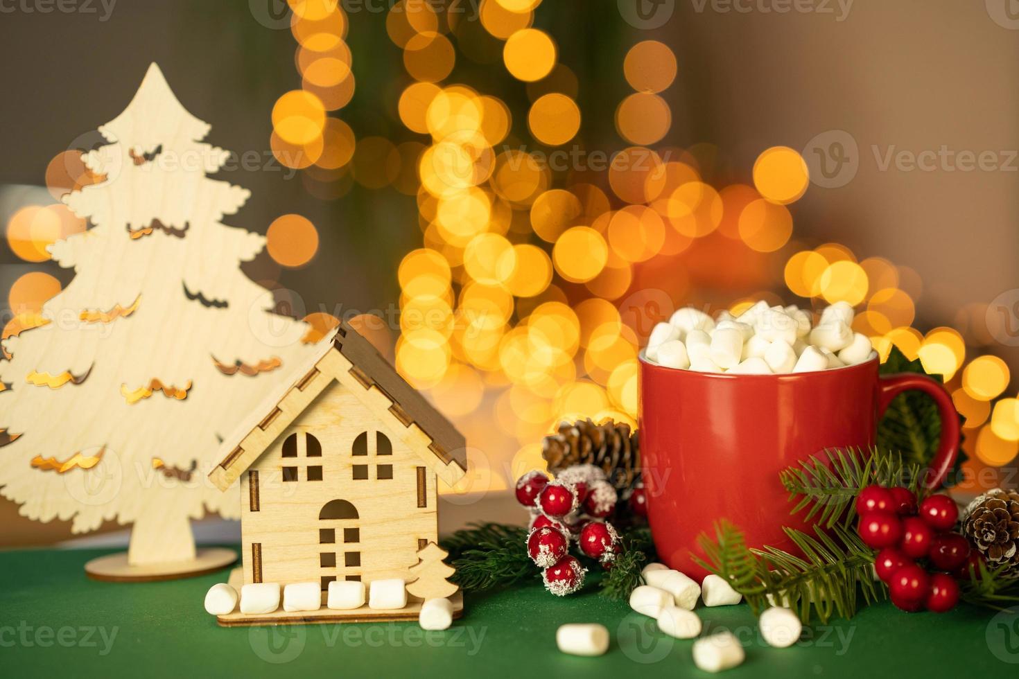 Christmas card with red mug of cocoa with marshmallow wooden house and fir tree and bokeh from garland light photo