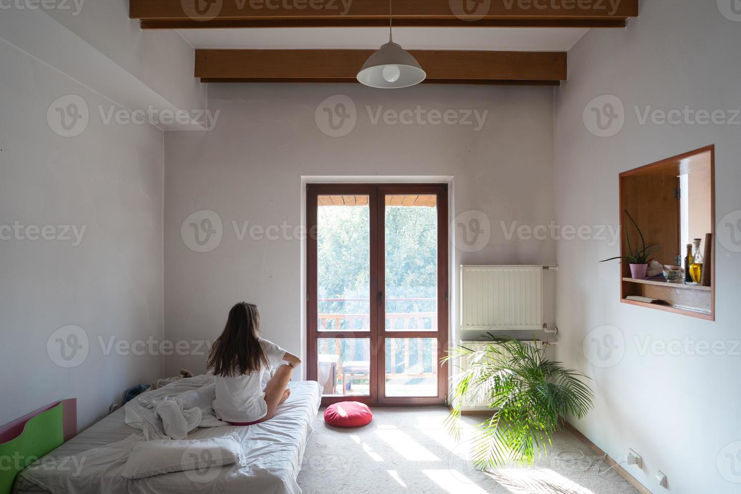 Young woman sitting on bed and looking through the window. photo