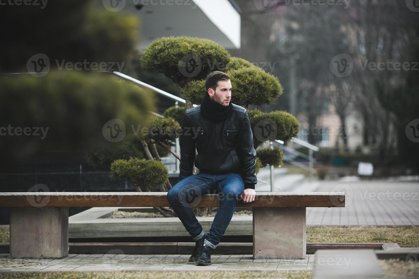A man dressed in jeans and black jacket seats on a bench photo