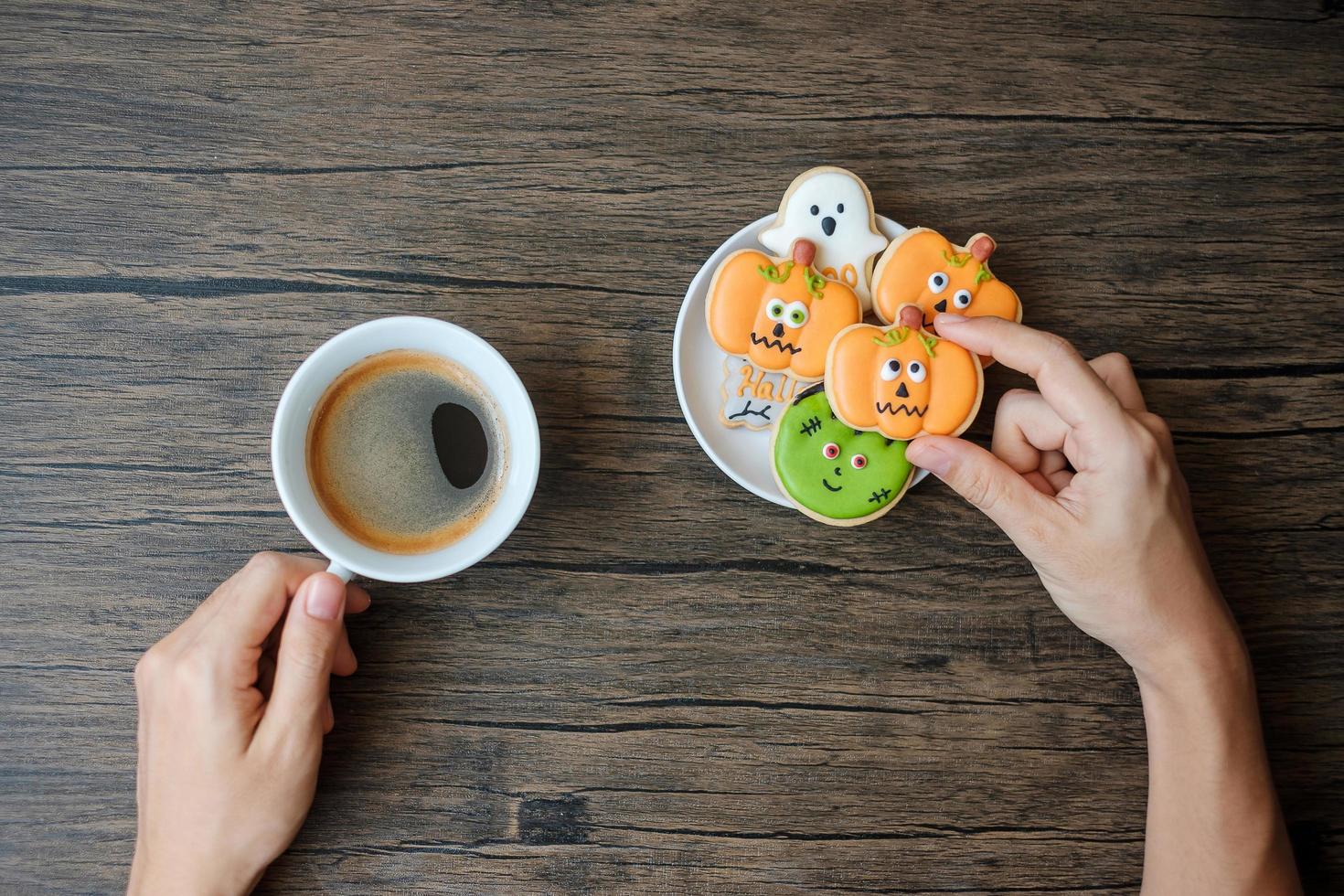 mano sosteniendo una taza de café mientras come divertidas galletas de halloween. feliz día de halloween, truco o amenaza, hola octubre, otoño, concepto tradicional, fiesta y vacaciones foto