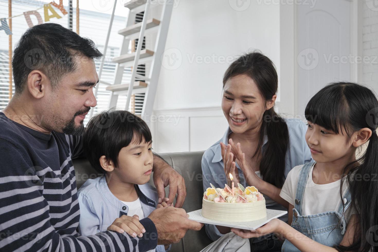 Happy Asian Thai family, young son is surprised with birthday cake, blows out candle, and celebrates joy party with parents and sister in living room together, wellbeing domestic home special event. photo