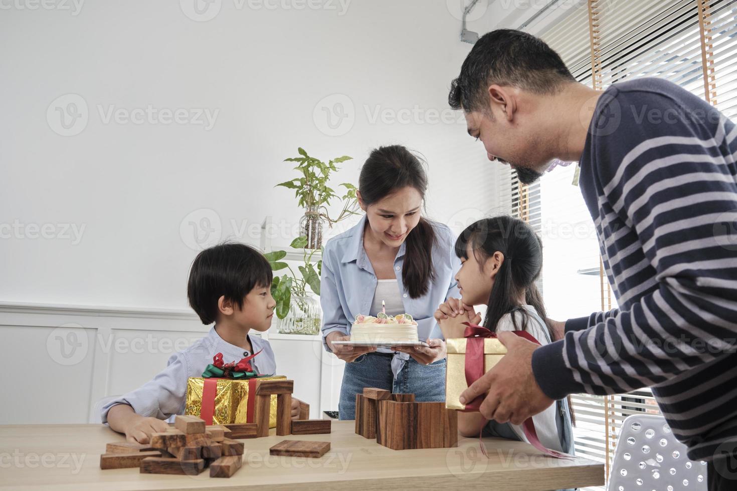 Happy Asian Thai family, young daughter is surprised with birthday cake and gift, blows candle, pray and cheerful celebrates party with parents together at dining table, domestic home event lifestyle. photo