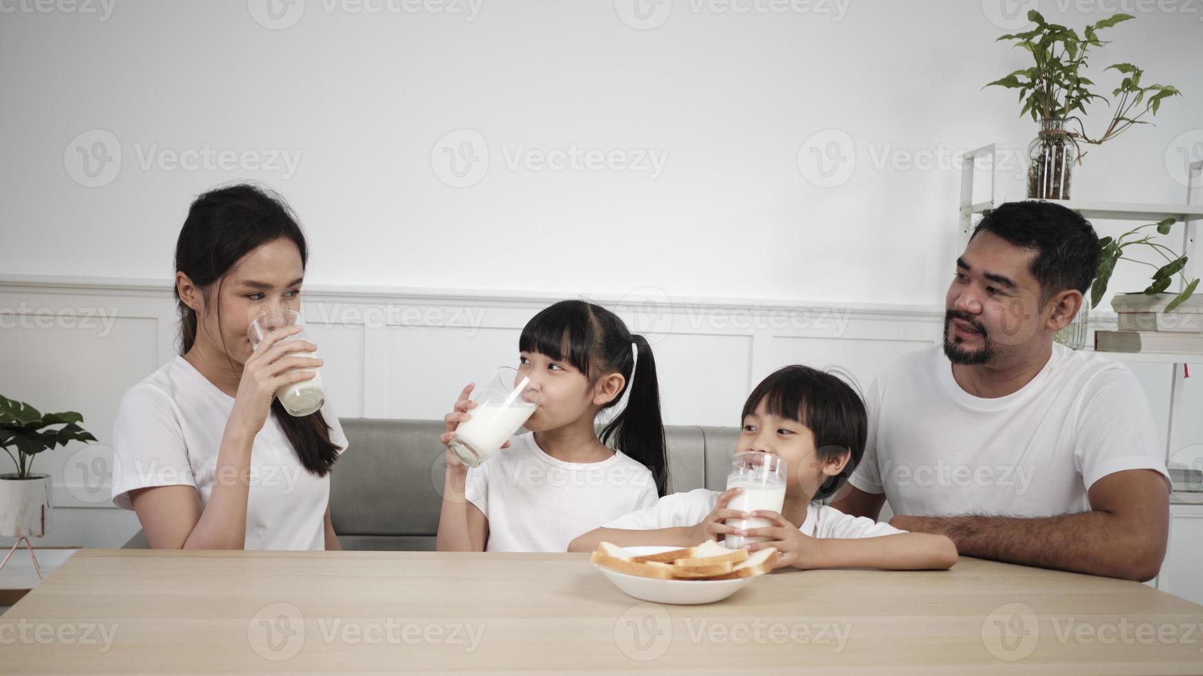 A Healthy Asian Thai family, little children, and young parents drink fresh white milk in glass and bread joy together at a dining table in morning, wellness nutrition home breakfast meal lifestyle. photo