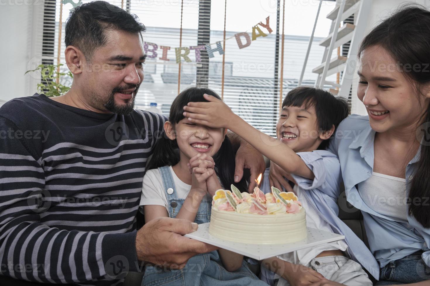 Happy Asian Thai family, young daughter is surprised with birthday cake, blows candle, pray and cheerful celebrates party with parents together in living room, wellbeing domestic home event lifestyle. photo