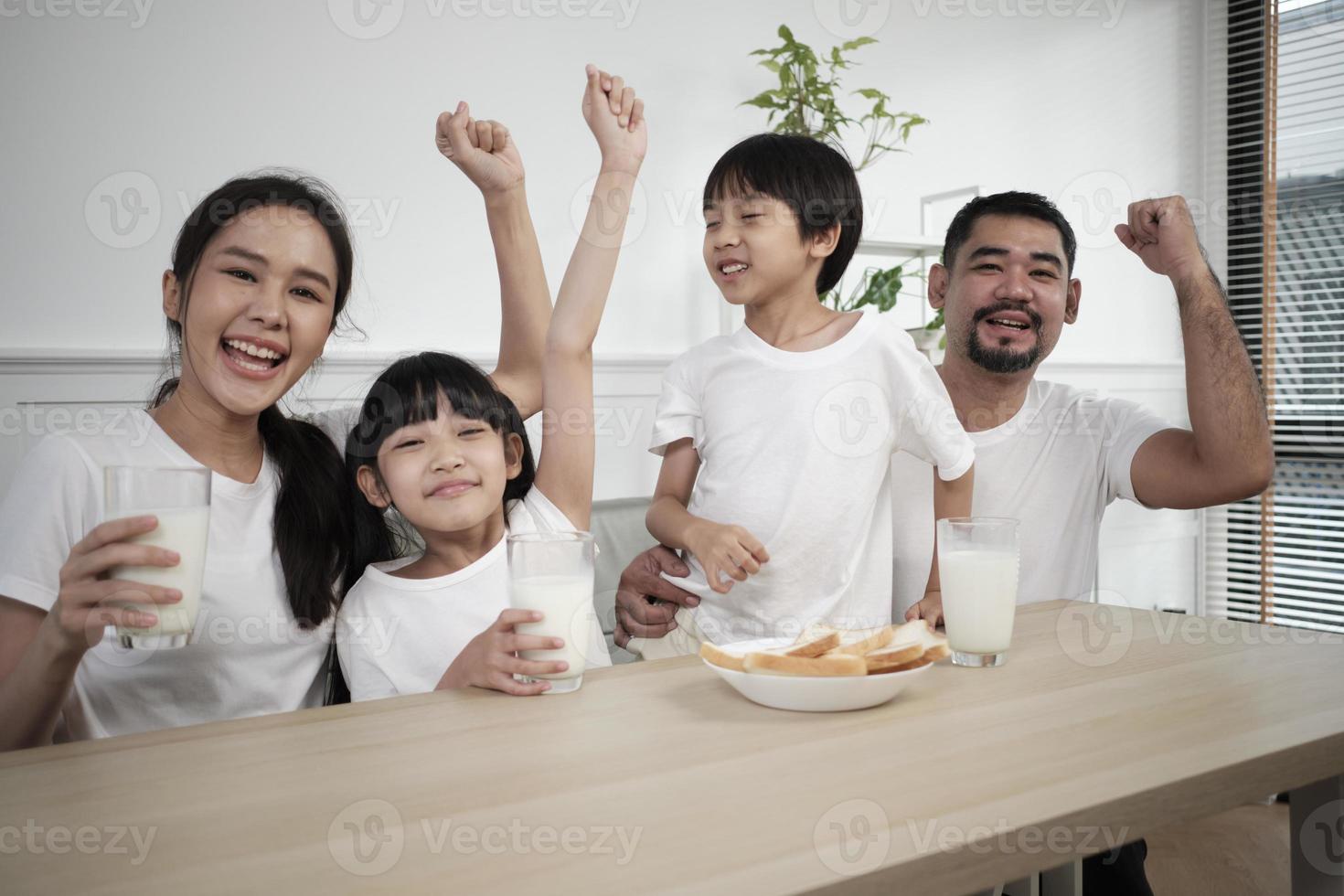 una familia tailandesa asiática saludable, niños pequeños y padres jóvenes beben leche blanca fresca en un vaso y pan de alegría juntos en una mesa de comedor en la mañana, bienestar nutrición hogar desayuno comida estilo de vida. foto