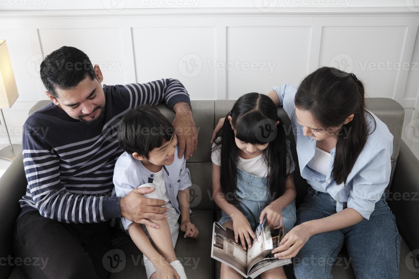 Asian Thai family, adult dad, mum, and children happiness home living relaxing activities and reading book together, leisure on sofa in white room house, lovely weekend, wellbeing domestic lifestyle. photo