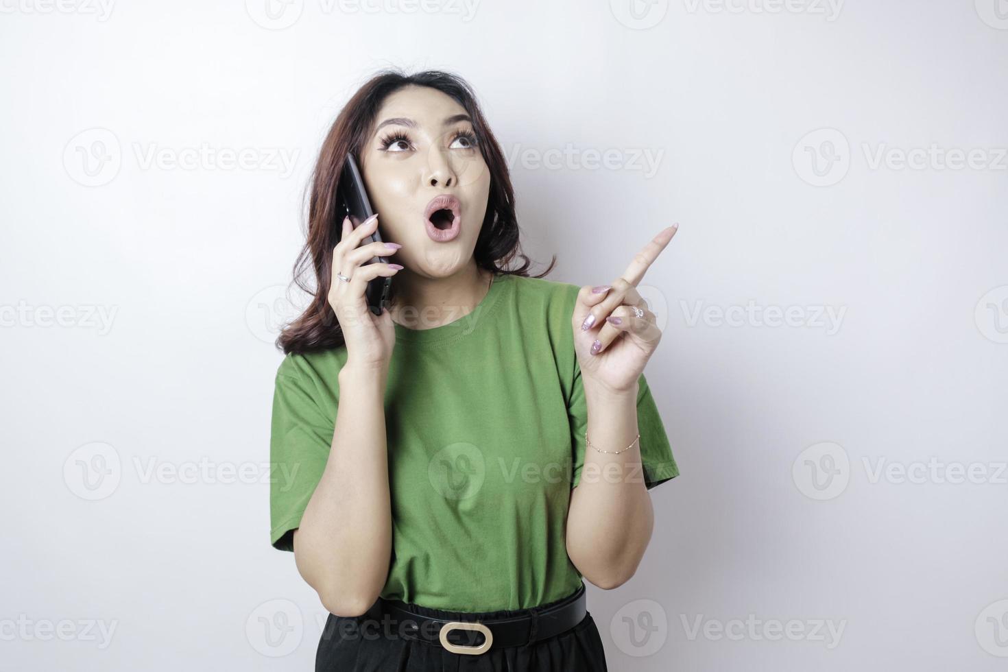 Shocked Asian woman wearing a green t-shirt pointing at the copy space upside her holding her phone, isolated by a white background photo