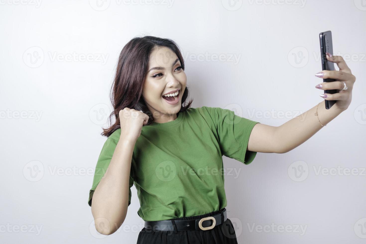 A young Asian woman with a happy successful expression wearing green t-shirt and holding smartphone isolated by white background photo