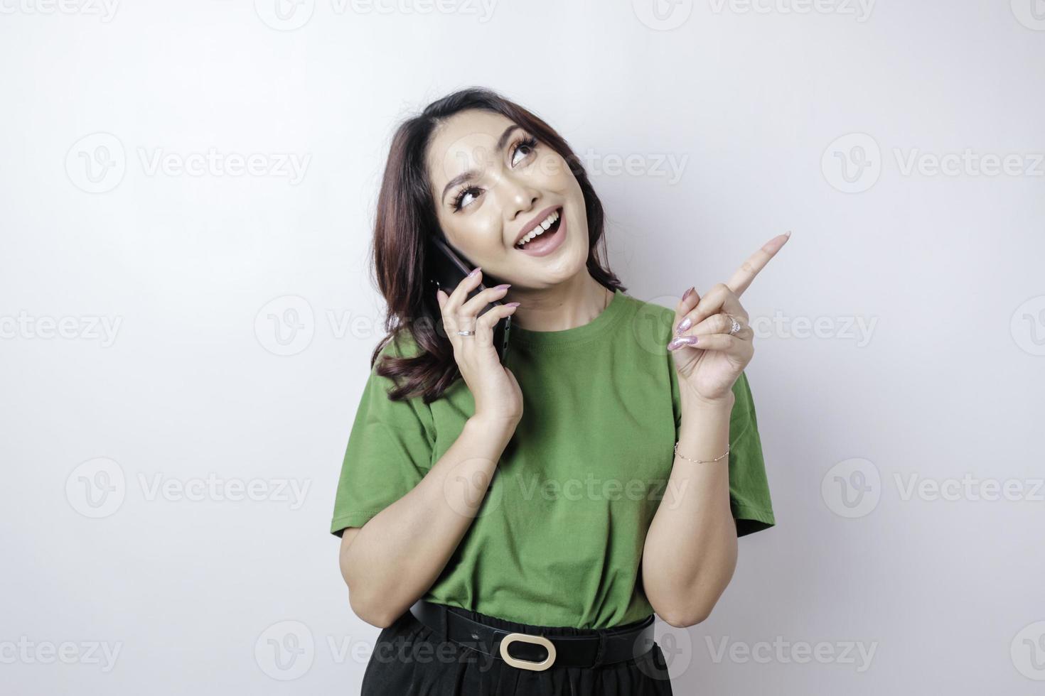 Smiling beautiful Asian woman pointing finger upward to empty space while holding her phone isolated white background photo