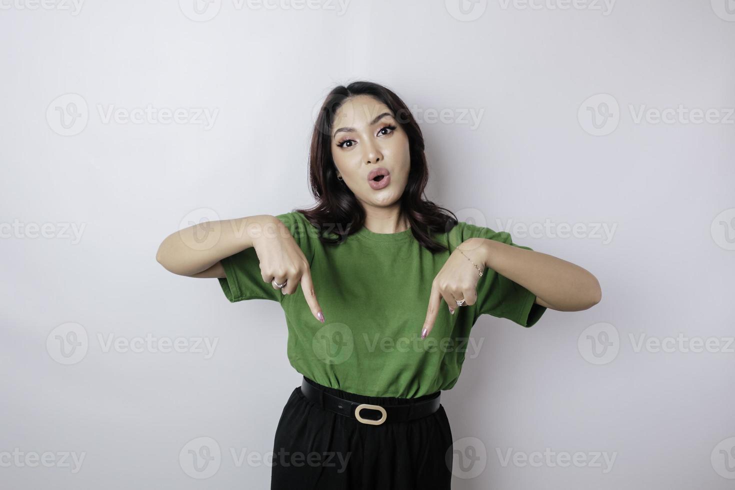 Shocked Asian woman wearing green t-shirt pointing at the copy space downside her, isolated by white background photo