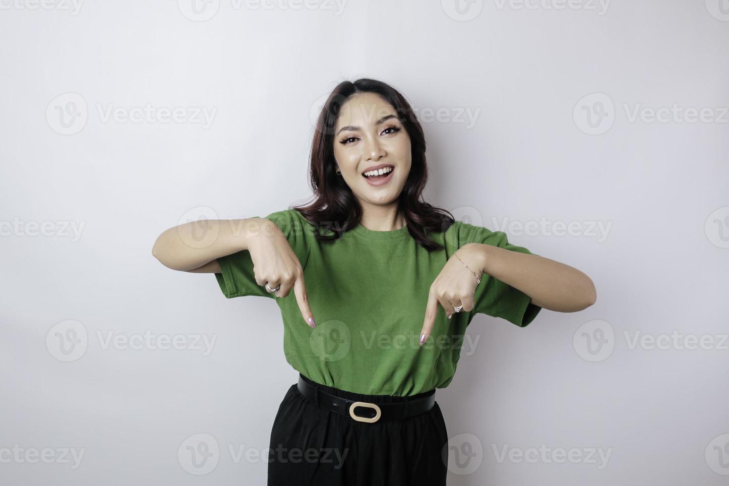 Excited Asian woman wearing green t-shirt pointing at the copy space downside her, isolated by white background photo
