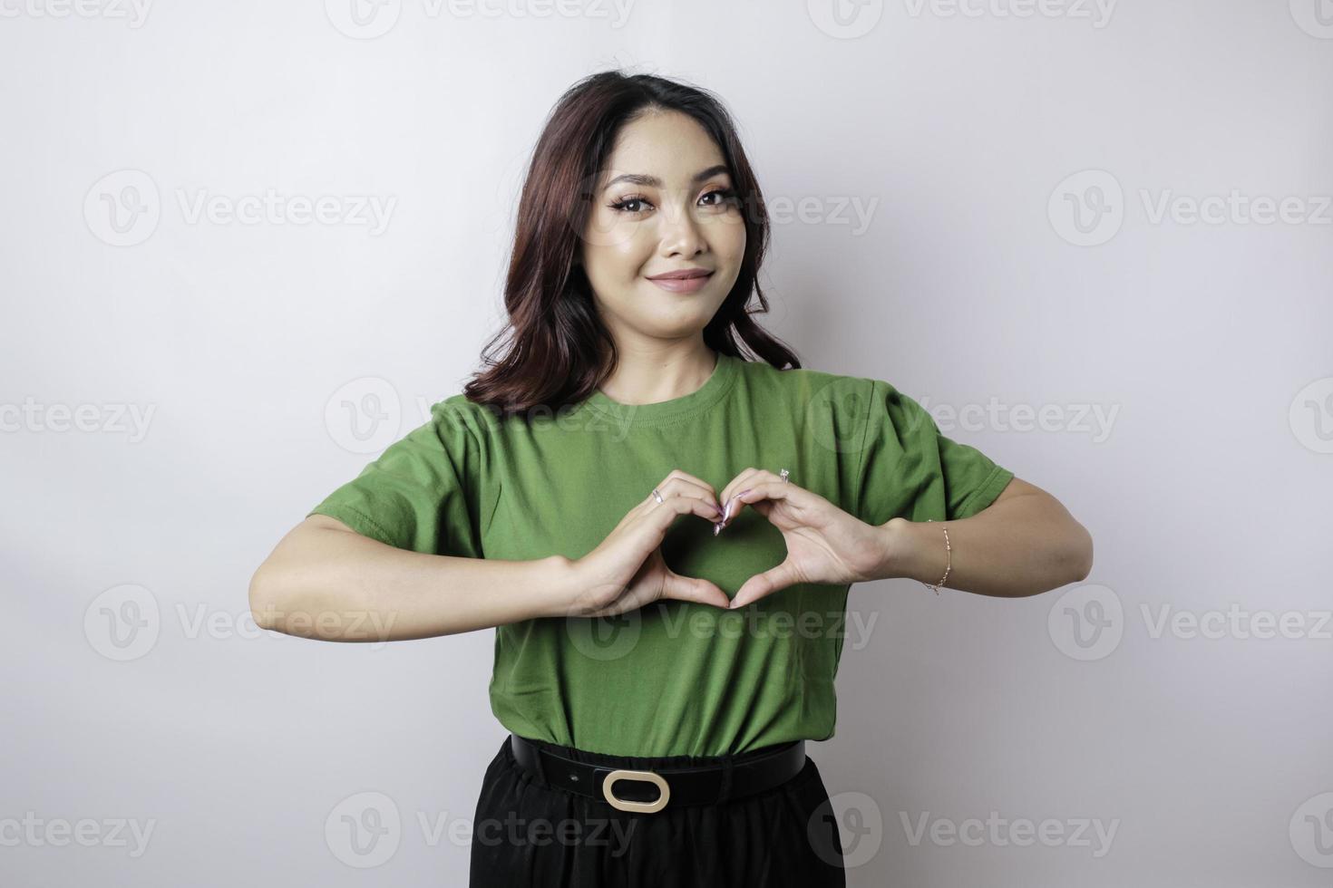 An attractive young Asian woman wearing a green t-shirt feels happy and a romantic shapes heart gesture expresses tender feelings photo