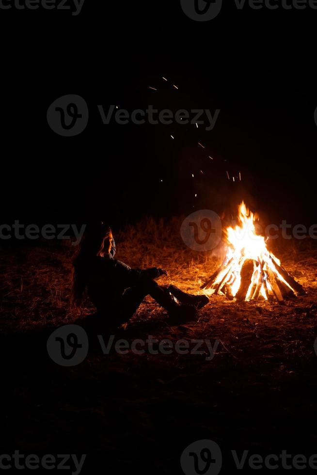 mujer sentada y calentándose cerca de la hoguera en el bosque nocturno. foto