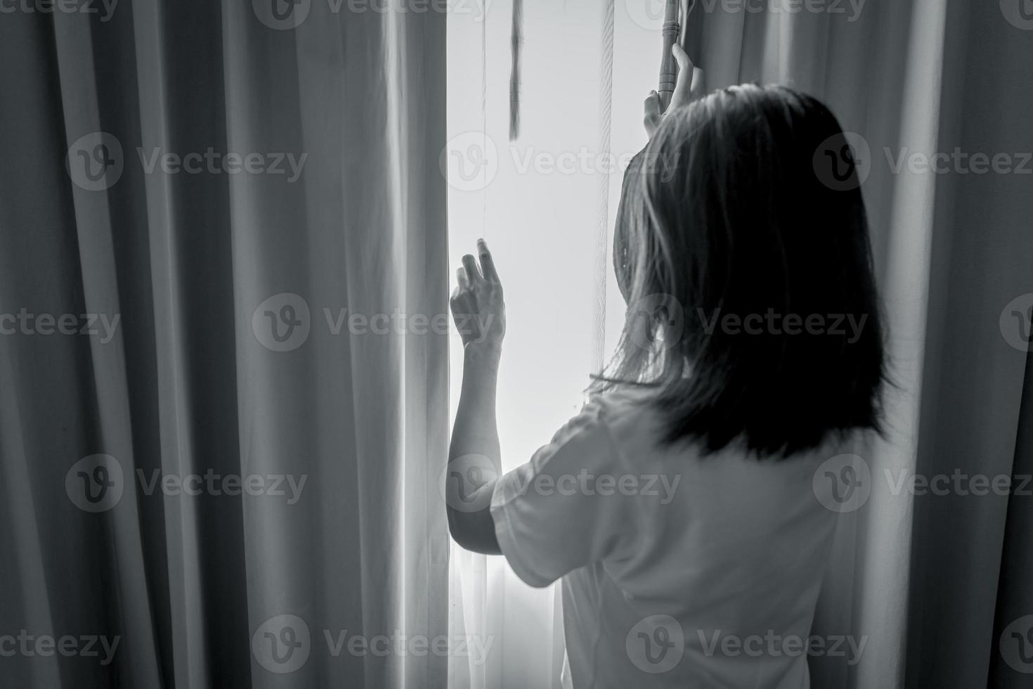Rear view of woman holding curtain and looking out of the bedroom. Depressed and stressed woman. World mental health day concept. Mental health awarenes. Thoughtful woman in dark room. Unhappy woman. photo