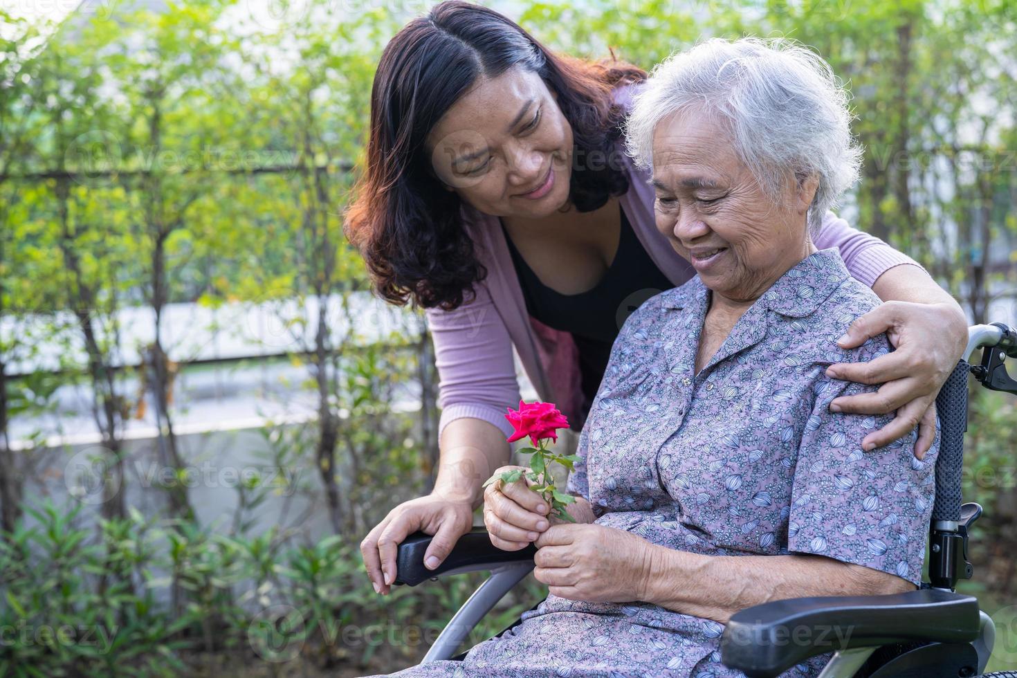 Caregiver daughter hug and help  Asian senior or elderly old lady woman holding red rose on wheelchair in park. photo