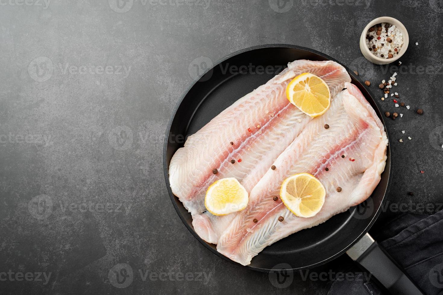 Raw pangasius fish fillet with lemon and spice in frying pan on concrete background photo