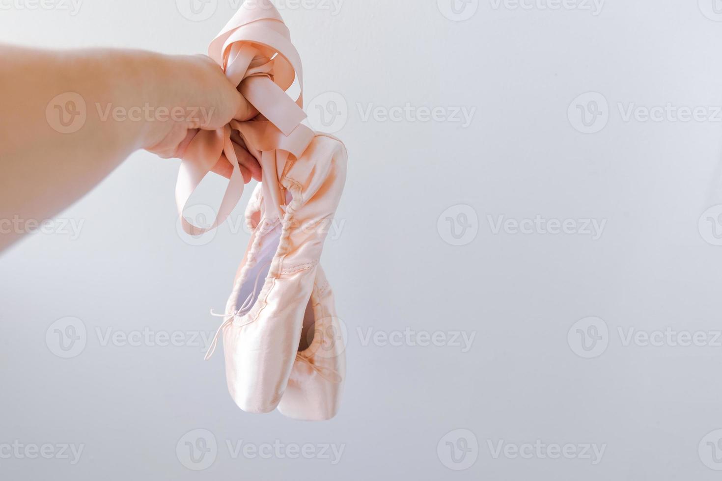 Ballerina dancer hand holding new pastel beige ballet shoes with satin ribbon isolated on white background. Ballerina classical pointe shoes for dance training. Ballet school concept. Copy space. photo