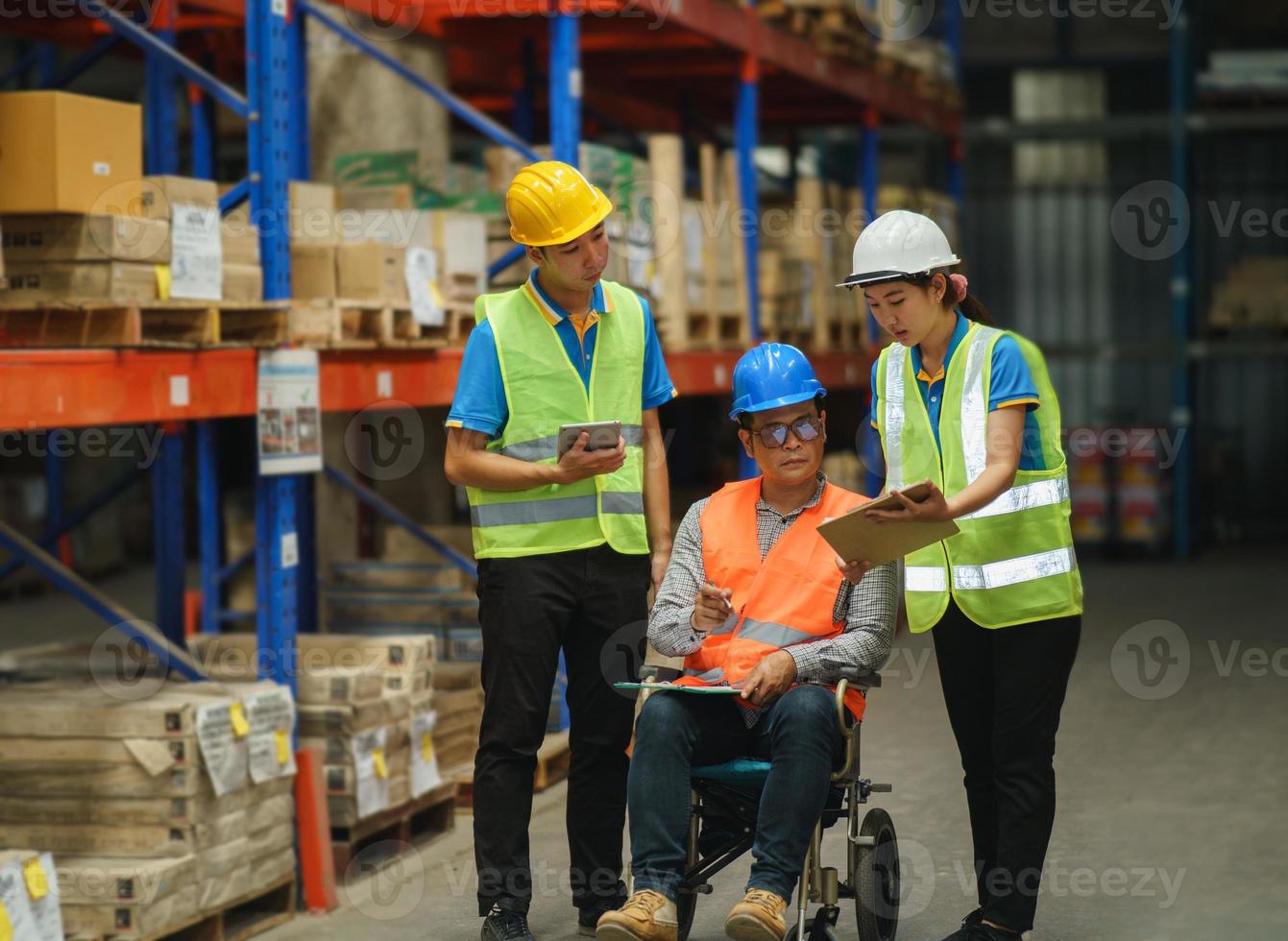 Manager handicapped in wheelchair man employee using tablet check stock work at warehouse. Worker wearing high visibility clothing and hard hat, helmet and checking count up goods boxes for delivery. photo