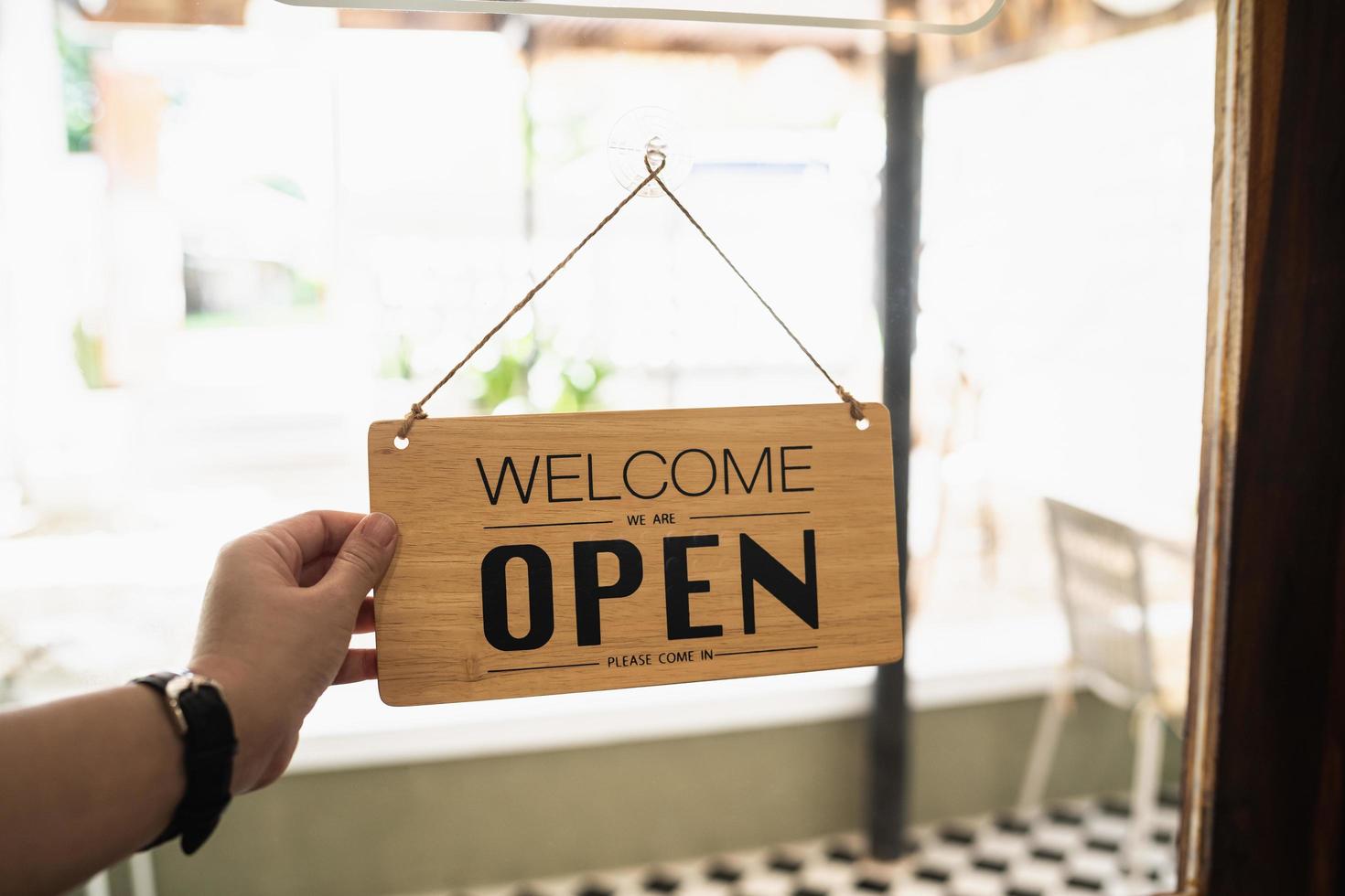 Hand woman hold open sign broad through the door glass. OPEN sign board through the glass of store window. text wooden board  door vintage shop sign opened store. photo
