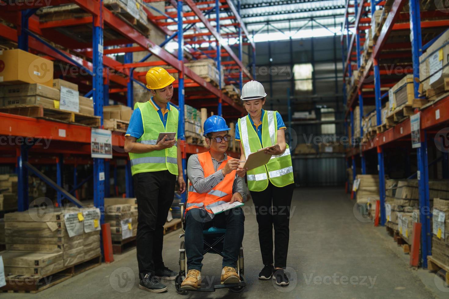 Manager handicapped in wheelchair man employee using tablet check stock work at warehouse. Worker wearing high visibility clothing and hard hat, helmet and checking count up goods boxes for delivery. photo