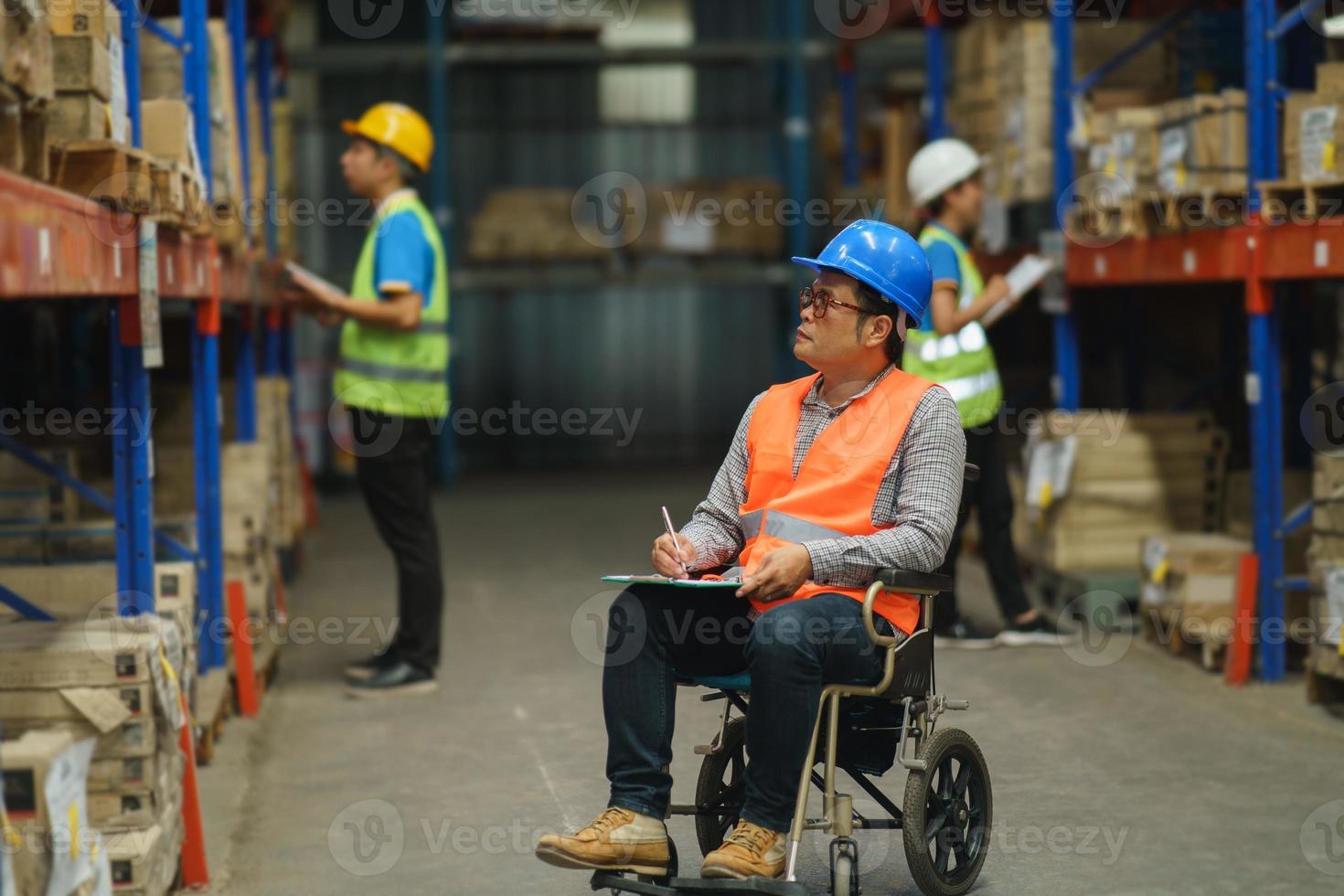 Manager handicapped in wheelchair man employee using tablet check stock work at warehouse. Worker wearing high visibility clothing and hard hat, helmet and checking count up goods boxes for delivery. photo