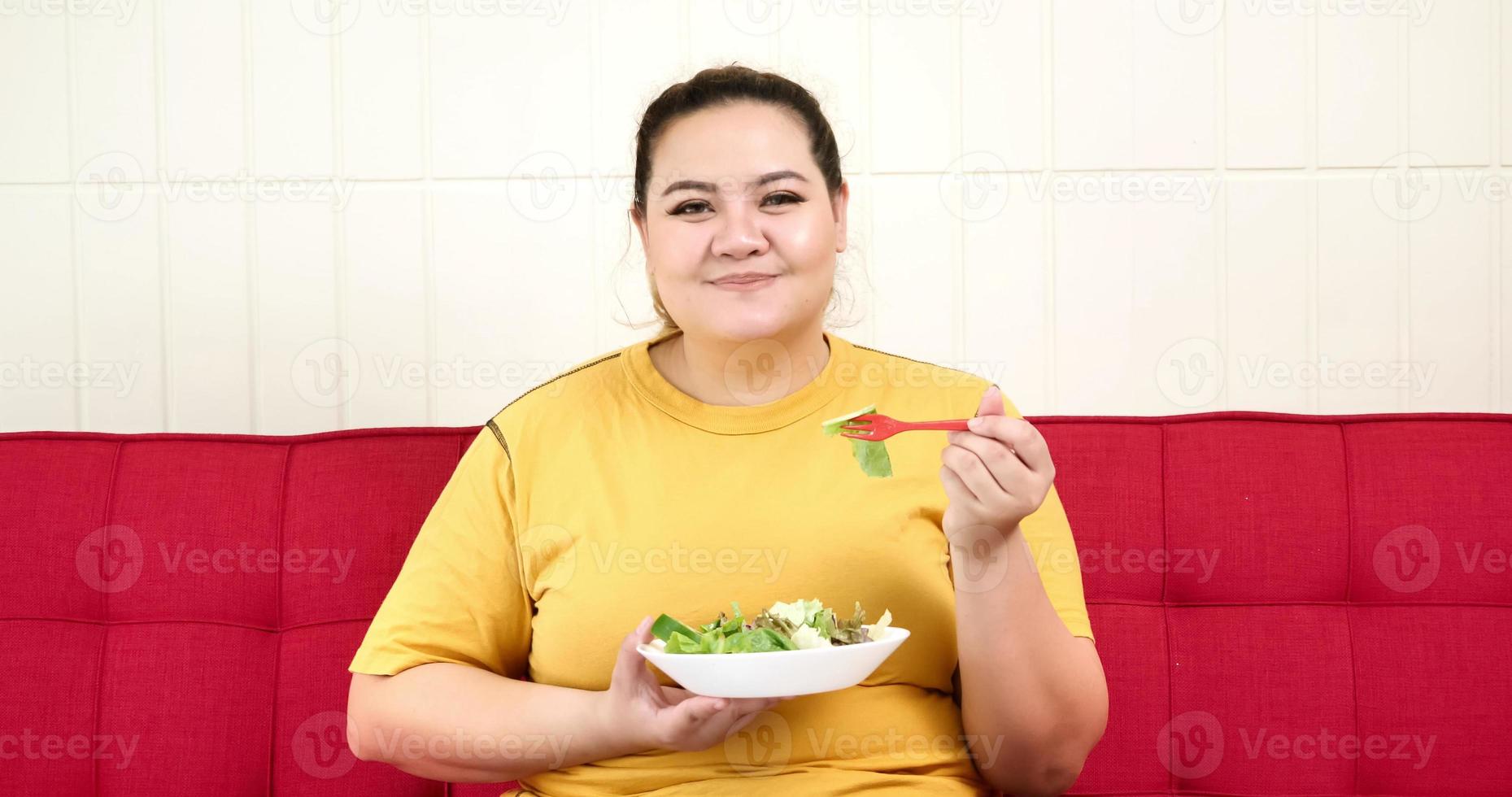 Chubby woman sitting on a sofa and eating salad. photo