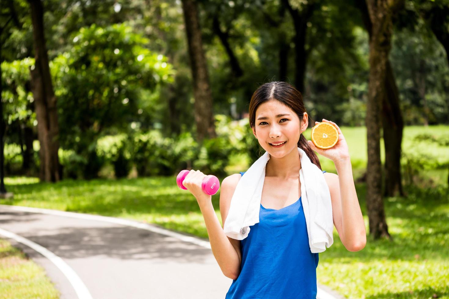 Portrait of attractive woman is holding dumbbell and orange in the park. photo