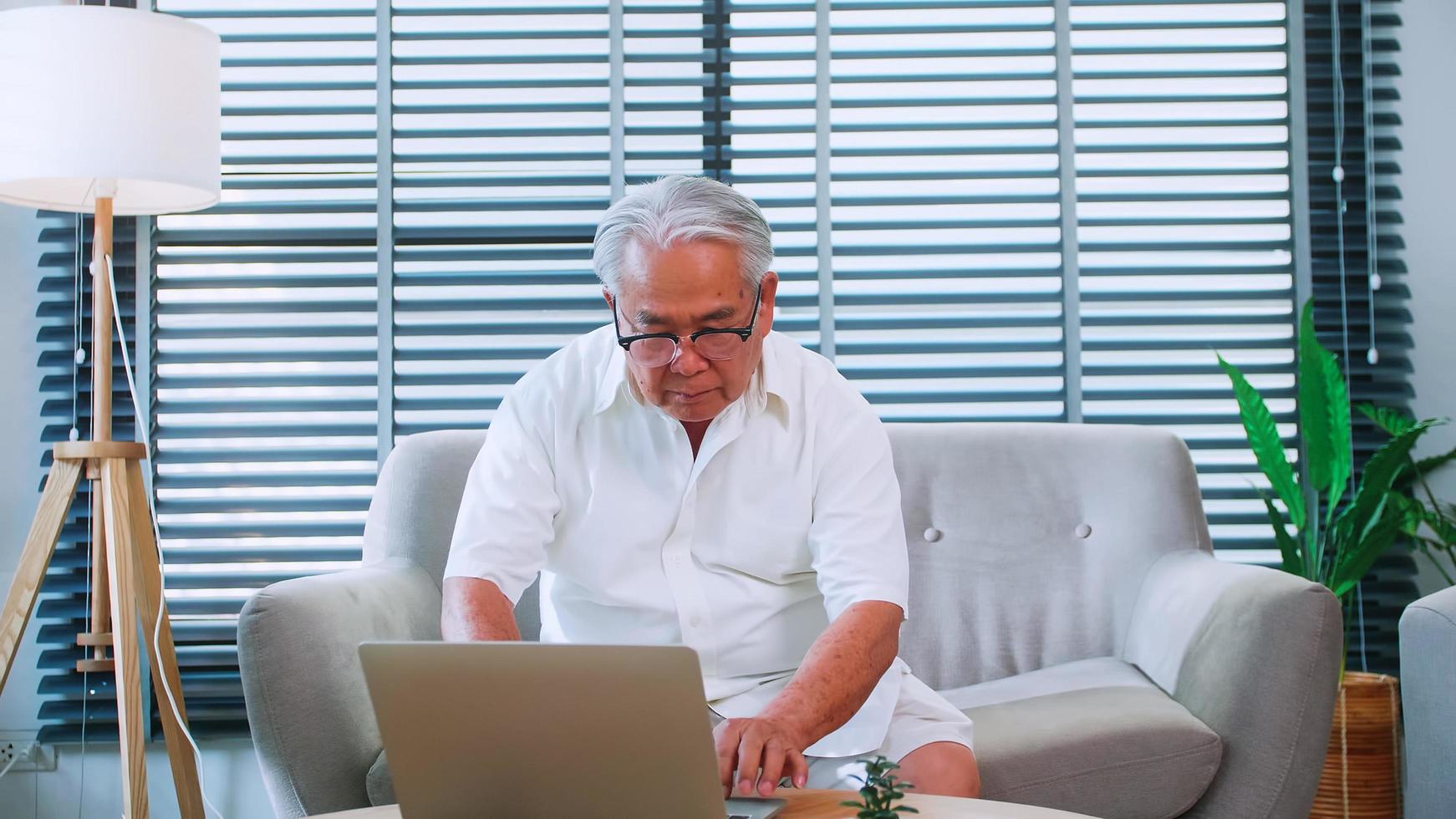 An elderly man reading news with the tablet on sofa at home. An old Asian man is searching for information on the Internet while sitting in living room photo