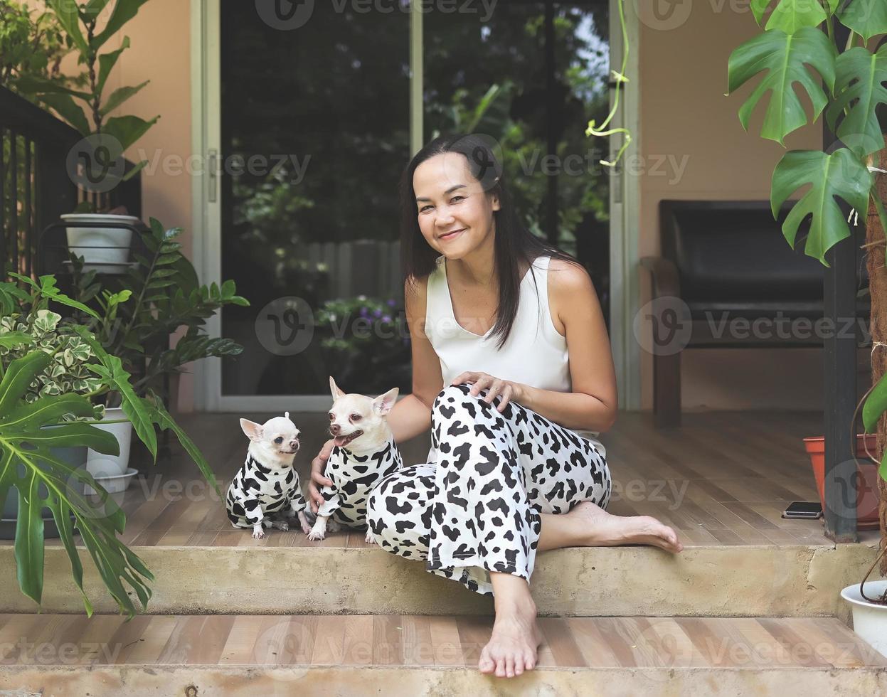 mujer asiática y sus dos perros chihuahua vestidos con un traje dálmata o de vaca sentados en un balcón con plantas de casa, sonriendo y mirando la cámara. foto