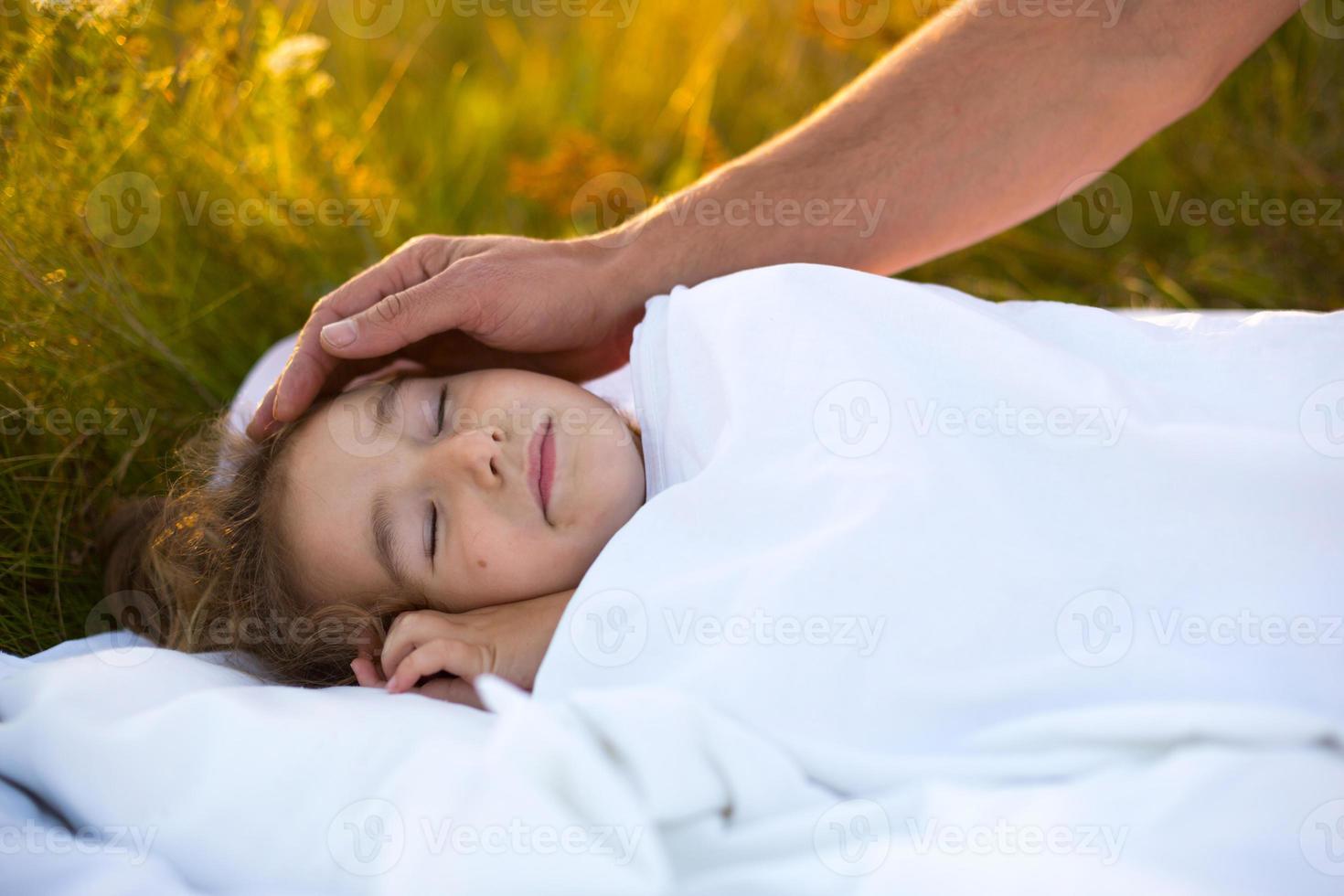 Girl sleeps on white bed in the grass, fresh air. Dad's hand gently pats his head. Care, protection, International Children's Day, mosquito bites photo