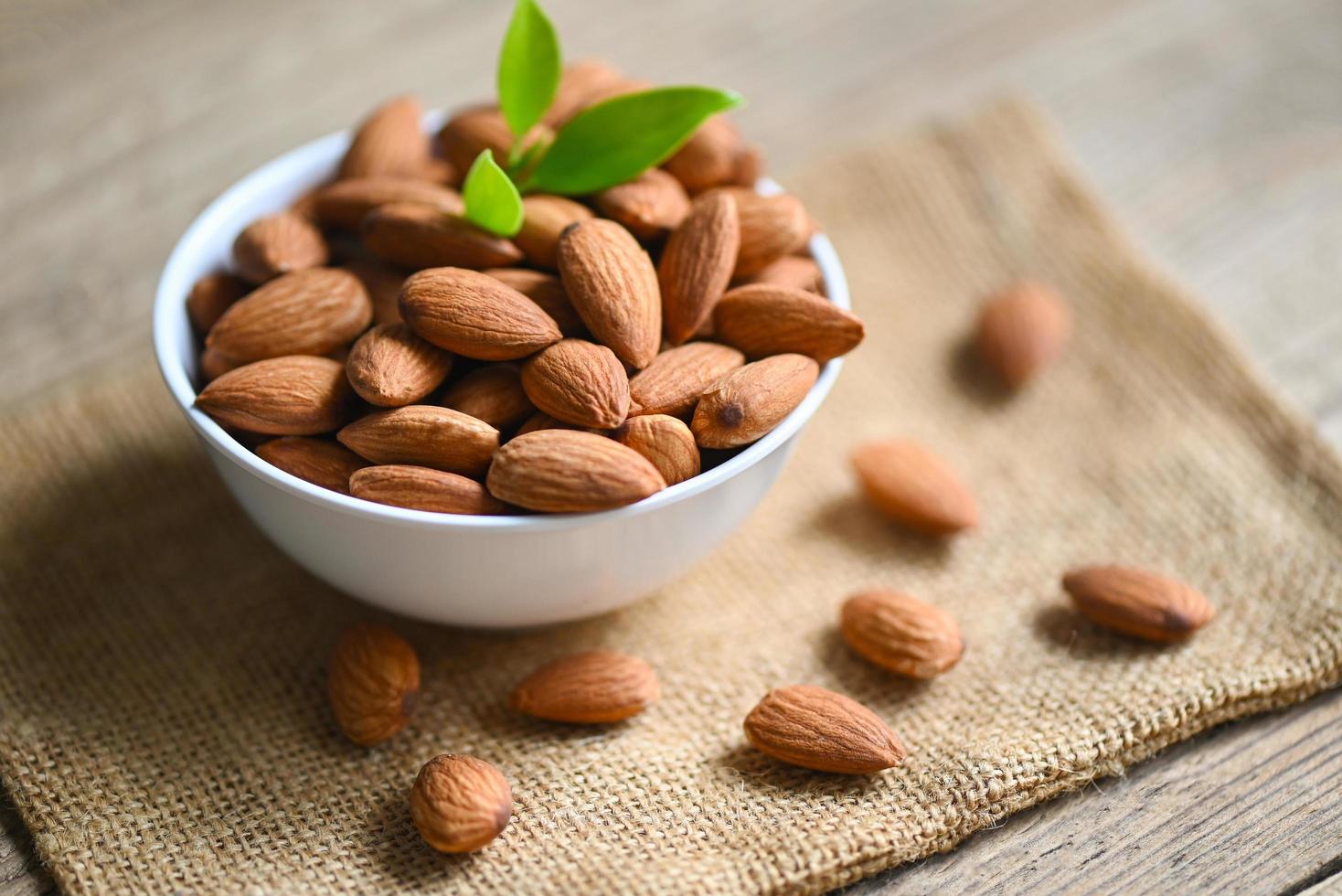 Almonds nuts on white bowl and green leaf on sack background, Delicious sweet almonds on the wooden table, roasted almond nut for healthy food and snack photo