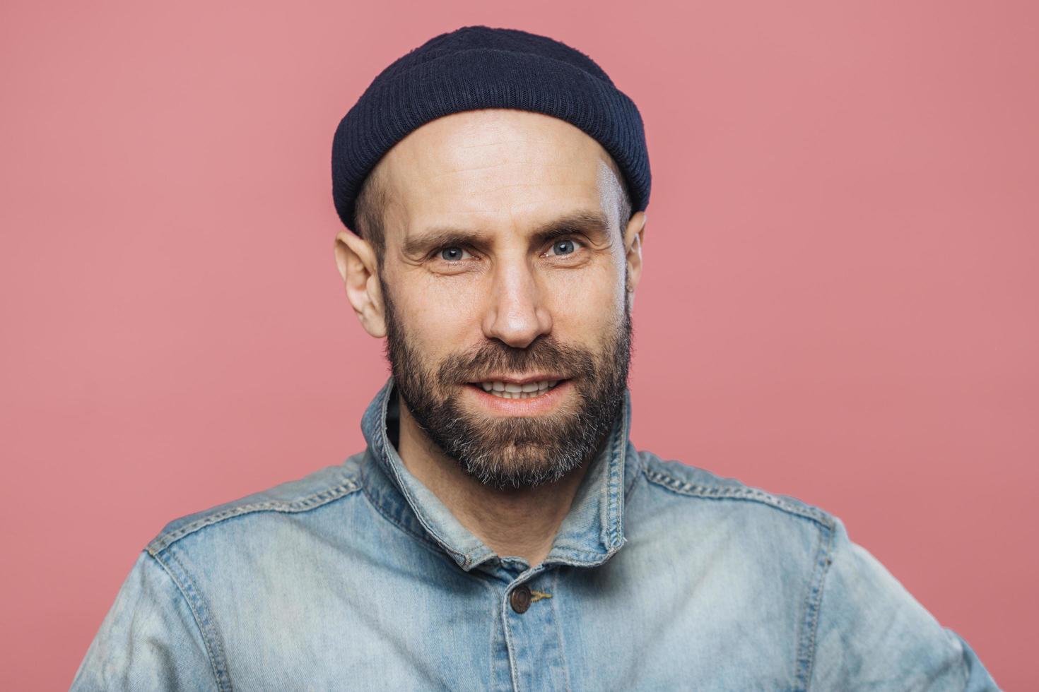 modelo masculino satisfecho con barba gruesa y bigote mira con expresión segura a la cámara, usa sombrero y chaqueta de moda, aislado sobre fondo rosa. personas, emociones, expresiones faciales foto