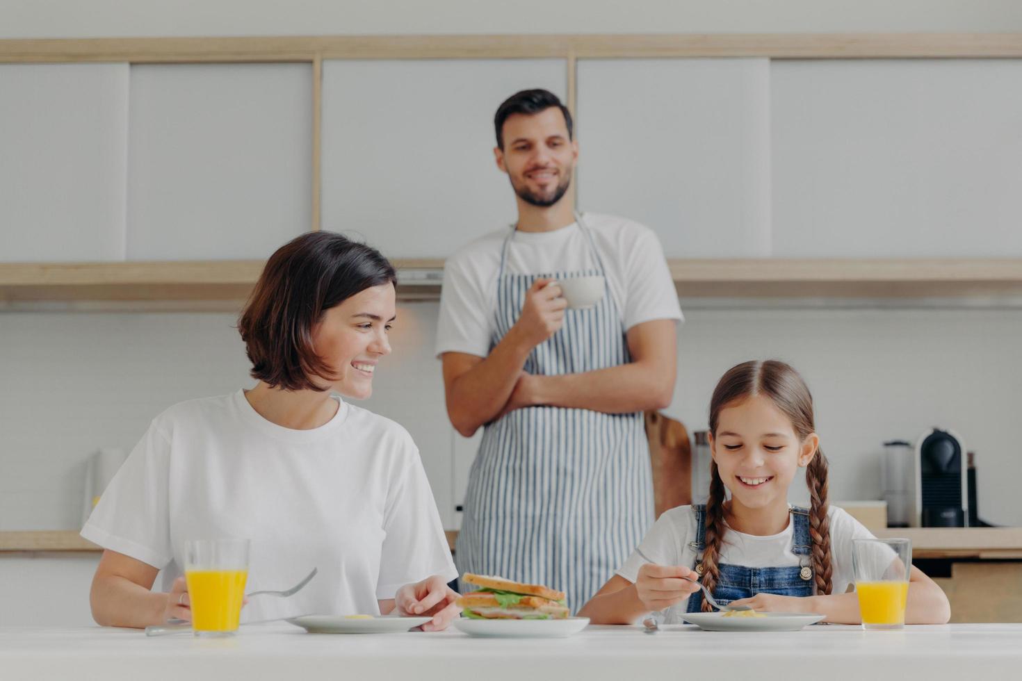 Happy mother talks to daughter while have breakfast. Father stands behind, prepared delicious dish for family. Friendly family members meet at kitchen during weekend, enjoy nice conversation photo